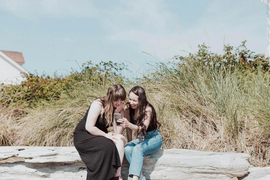 Zwei Frauen sitzen lachend auf einem Baumstamm am Strand und schauen auf ein Handy.